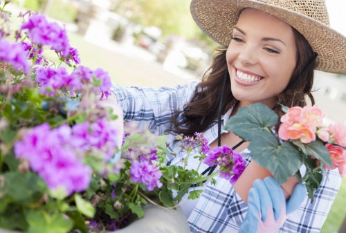 A woman with a broad smile, wearing a wide-brimmed straw hat and blue gardening gloves, is engaged in planting or tending to flowering plants in a well-maintained outdoor garden. In the foreground, vibrant purple and pink flowering plants are arranged in containers or flower beds, showcasing a variety of lush, healthy foliage. The garden features a neatly mowed lawn with dense, green grass that extends into the background, bordered by a hedge or shrub border. There are pathways or paved areas partially visible on the sides, and the overall environment suggests a sunny day with natural light illuminating the scene. The setting is a private garden space, possibly a front or back garden in Holborn, that benefits from professional gardening and lawn care services, such as those offered by Lawn Mowing Holborn, to maintain its tidy and inviting appearance. The image emphasizes the natural beauty and careful landscaping characteristic of a well-kept outdoor area in an urban residential context.